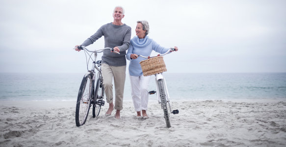 Picture of a later aged couple who are pushing bicycles on the sand.